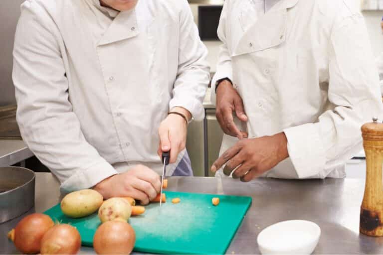 Two chefs in white coats stand over a cutting board while one demonstrates proper cutting technique on a carrot