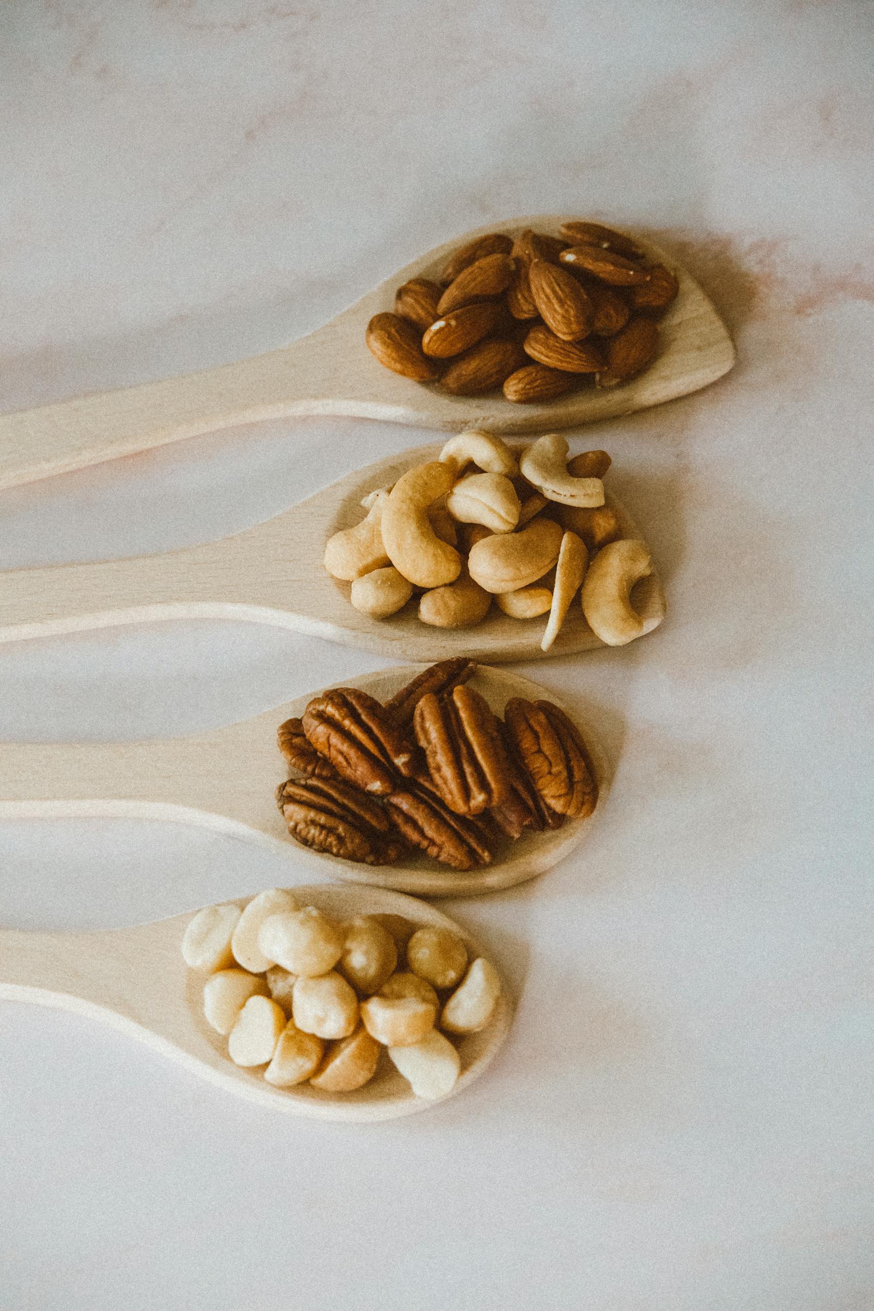 A vertical shot showcasing four types of nuts in wooden spoons, laid on a marble surface.