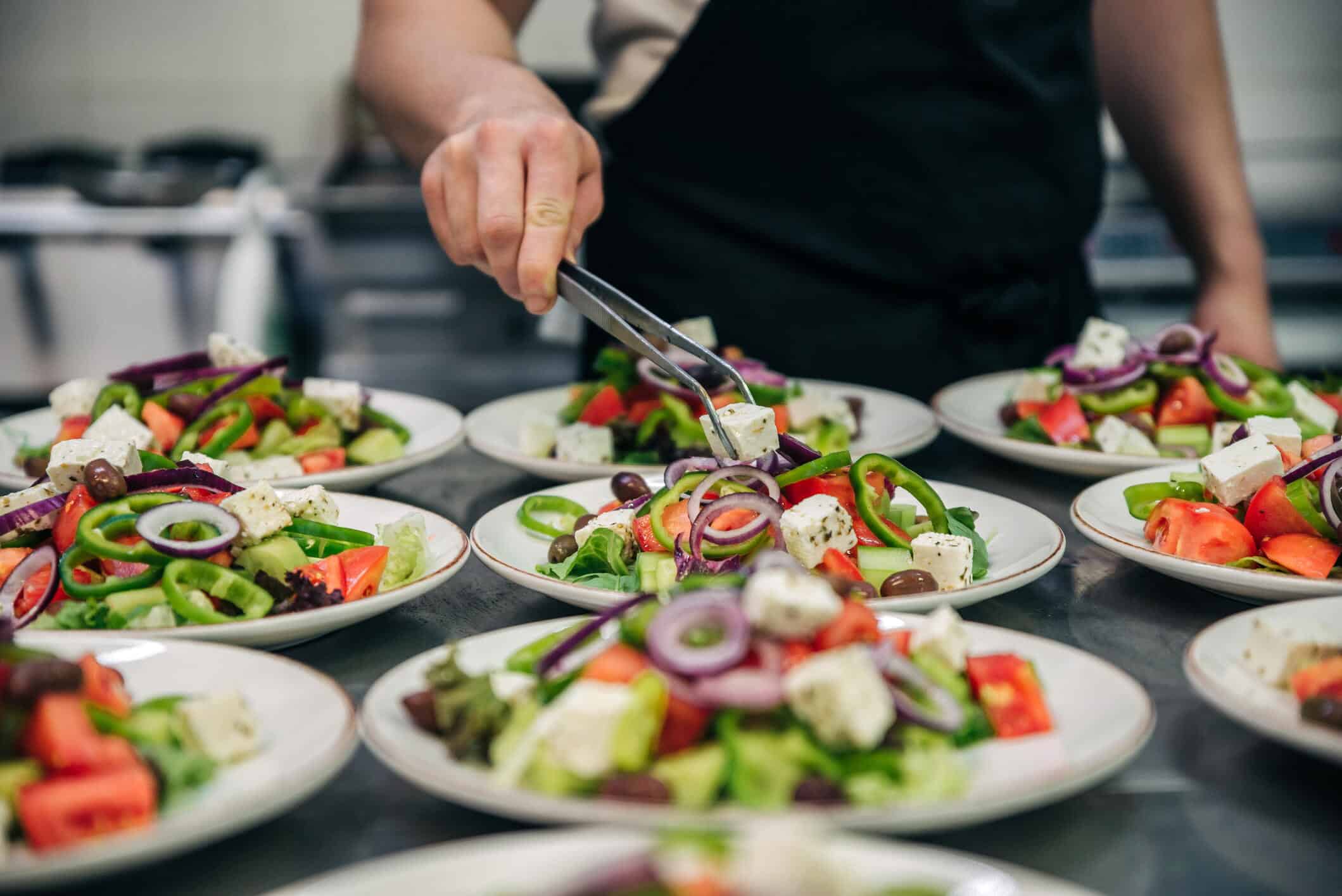 Chef finishing healthy greek salad on white plate with tweezers. almost ready to serve it on a table. Restaurant kitchen