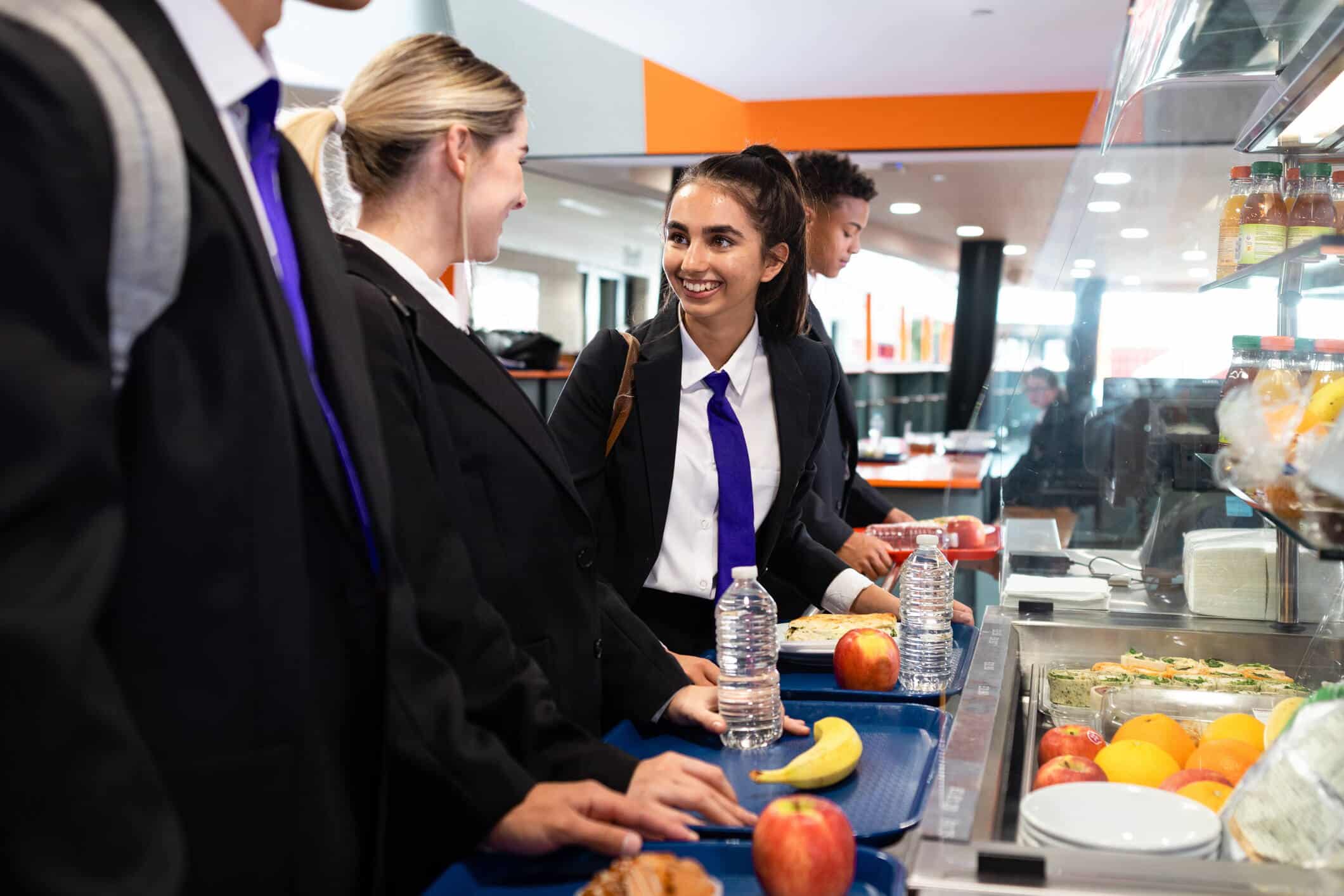 A shot of teenage students waiting in line together at the cafeteria in a school. The students are wearing school uniforms and waiting with their dinner trays.