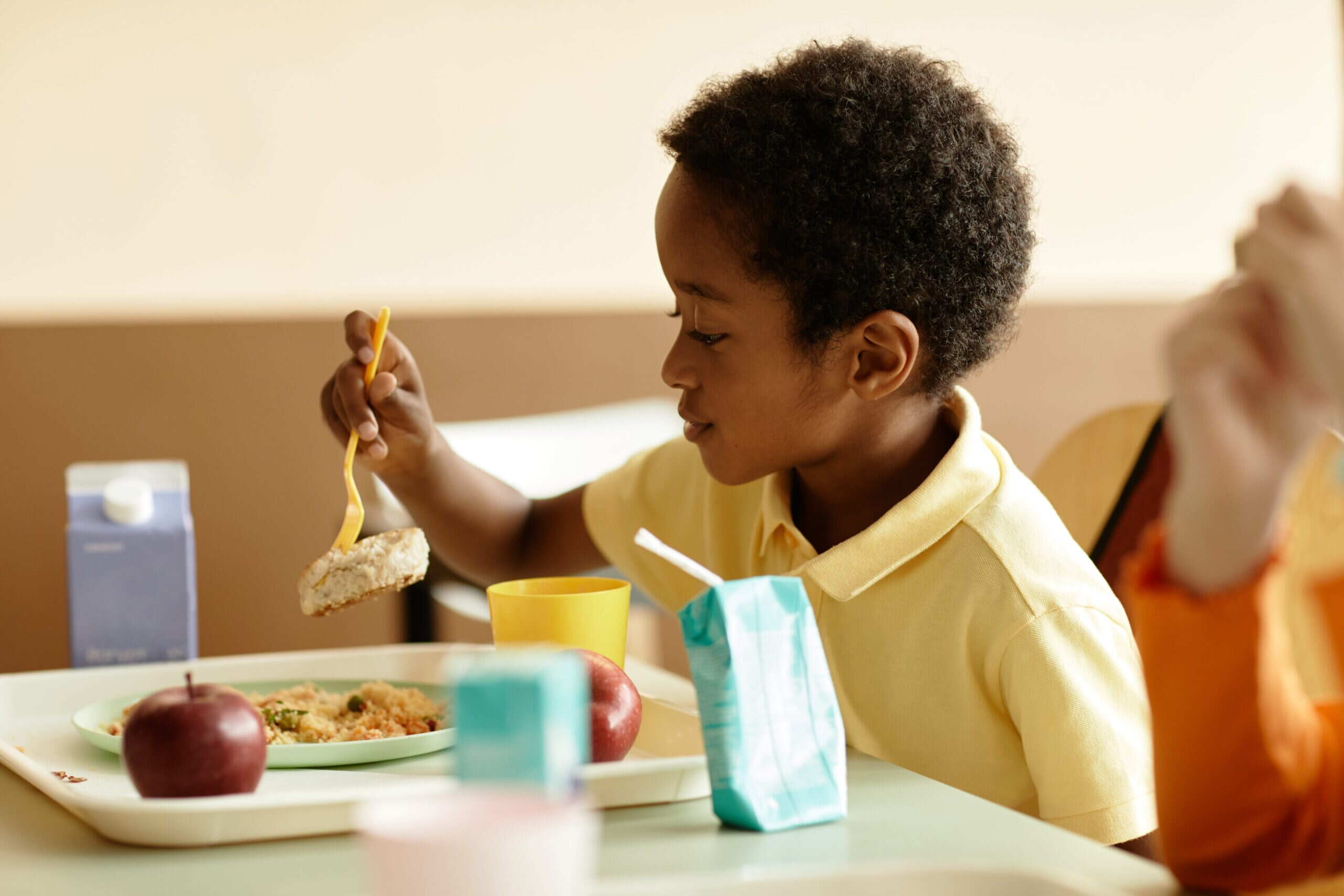 A child enjoying lunch at a school cafeteria, surrounded by tables and classmates.