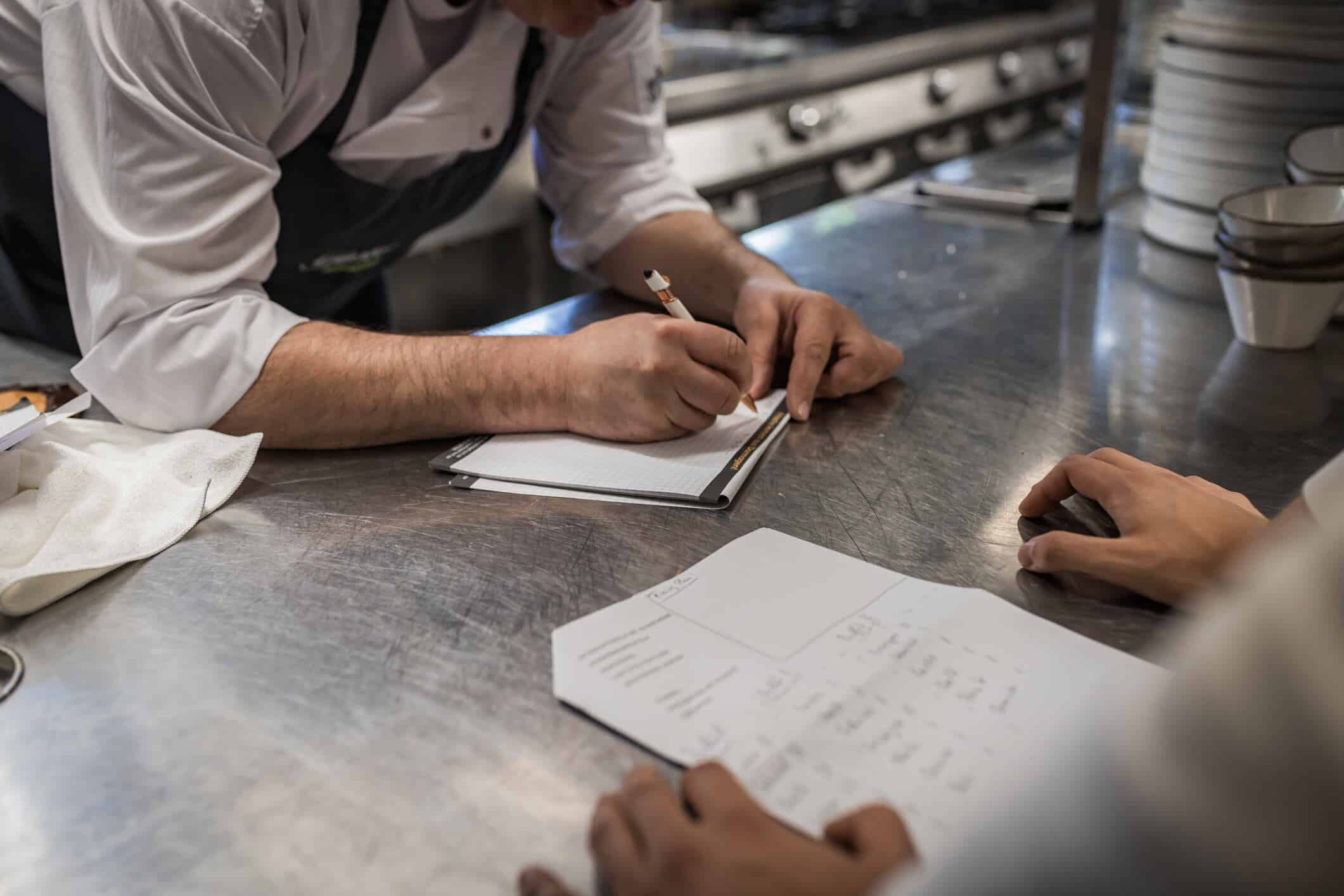 Two chefs work together in a busy professional kitchen, focused on menu planning and collaboration. The environment highlights teamwork and culinary preparation in a stainless steel setting.