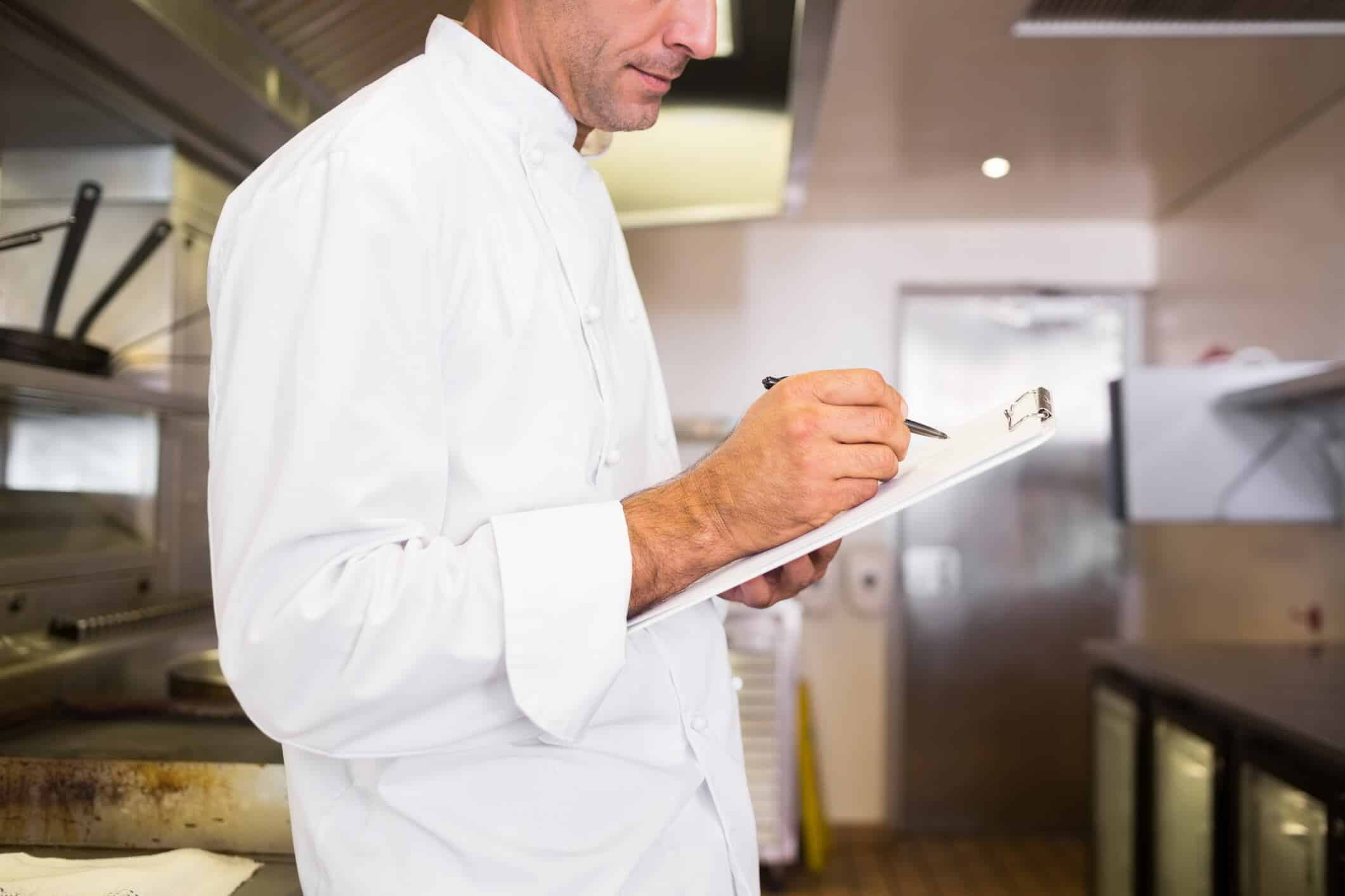 Side view of a concentrated male cook writing on clipboard in the kitchen